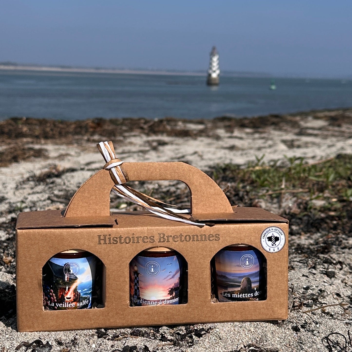 Coffret bougies artisanales Bretagne posé sur plage avec phare en arrière-plan