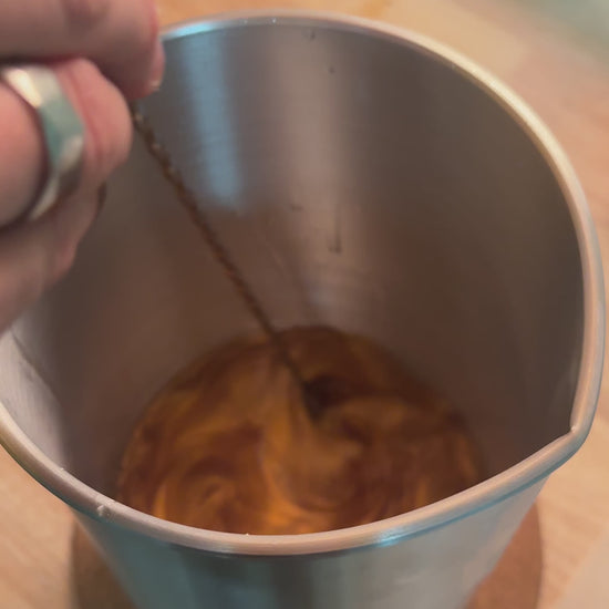 Cire fondue ambrée remuée avec une tige métallique dans un pichet en inox, vue de dessus, sur un plan de travail en bois.