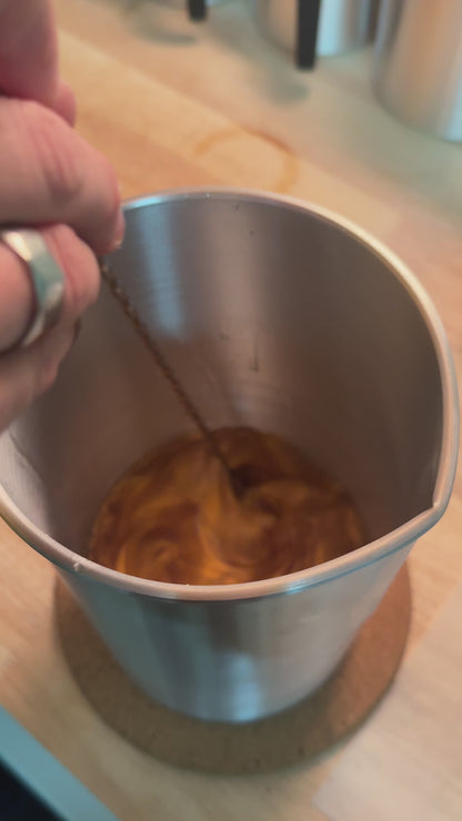 Cire fondue ambrée remuée avec une tige métallique dans un pichet en inox, vue de dessus, sur un plan de travail en bois.